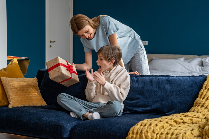 Young woman loving mother surprising happy excited son with birthday gift at home